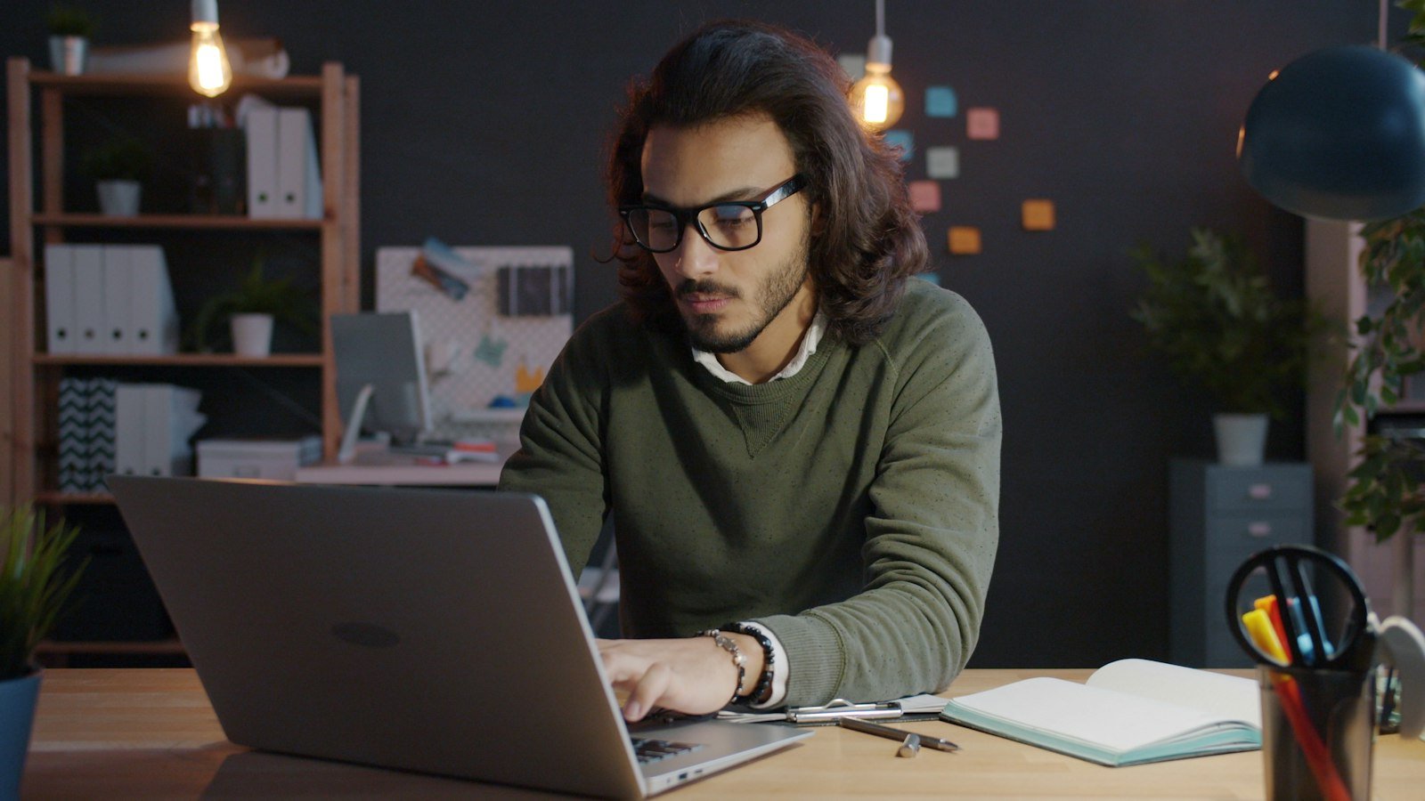 Freelancer in glasses working on a laptop and taking notes in an office at night